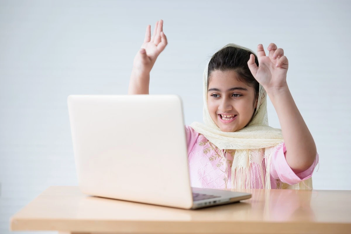Young Muslim girl raising both hands during an online lesson on a laptop