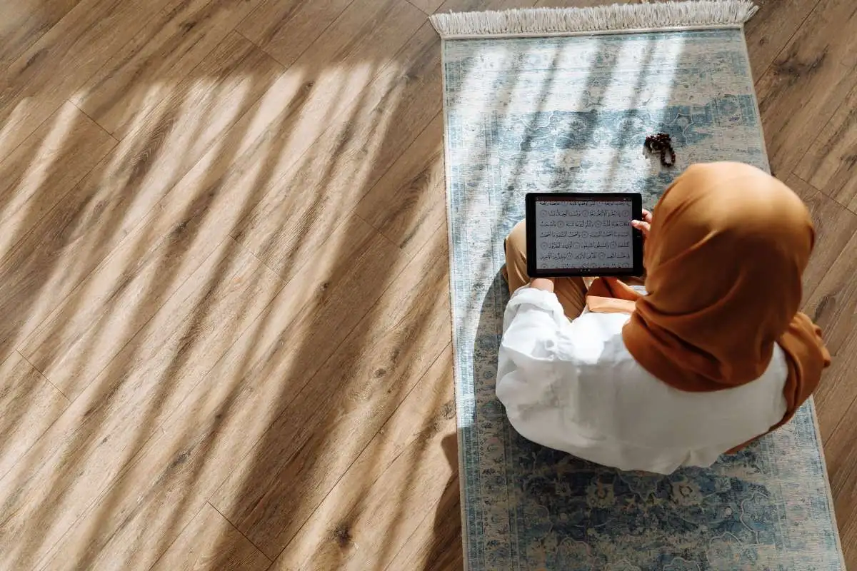 Woman reading the Quran on a tablet while seated on a prayer mat at home