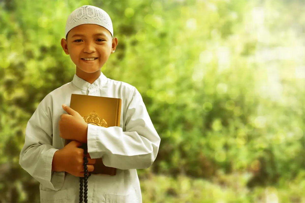 Smiling boy in a white thobe holding a Quran and prayer beads