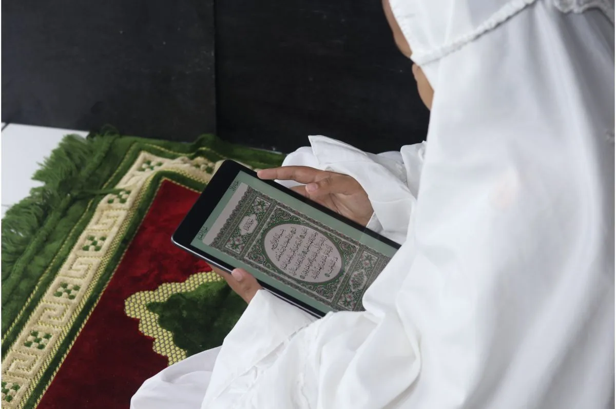 Person reading the Quran on a tablet beside a prayer mat